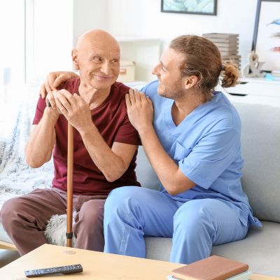 Ein Mann mit Gehstock sitzt auf einer Couch, während ein Krankenpfleger ihm zur Seite steht und ihn umsorgt.